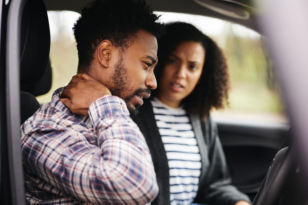 A man sitting in a car holds his neck in pain, while a concerned woman sits beside him, possibly after a car accident - Chiropractor Kennesaw GA A man sitting in a car holds his neck in pain, while a concerned woman sits beside him, possibly after a car accident - Chiropractor Kennesaw GA