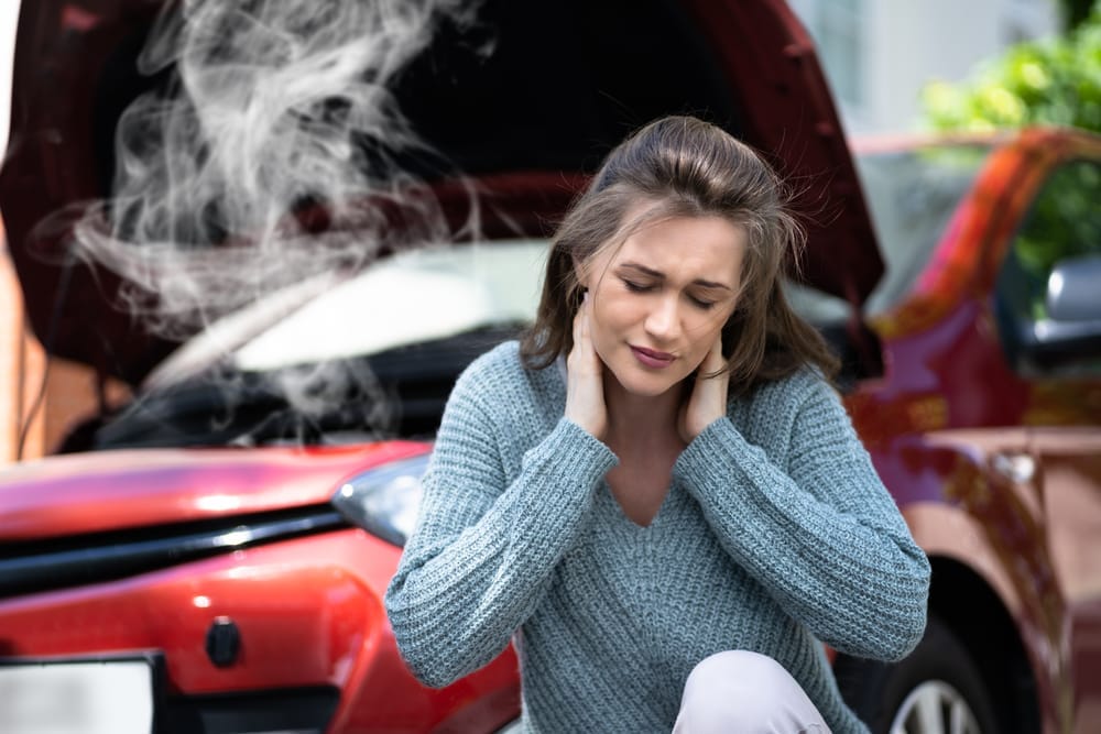 A woman sits in front of a car with an open hood and smoke rising, holding her neck in discomfort, possibly from an accident - Chiropractor Kennesaw GA A woman sits in front of a car with an open hood and smoke rising, holding her neck in discomfort, possibly from an accident - Chiropractor Kennesaw GA