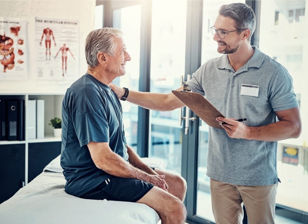 A chiropractor smiles while speaking with an older patient sitting on an examination table in a bright, modern clinic - Chiropractor Kennesaw GA A chiropractor smiles while speaking with an older patient sitting on an examination table in a bright, modern clinic - Chiropractor Kennesaw GA