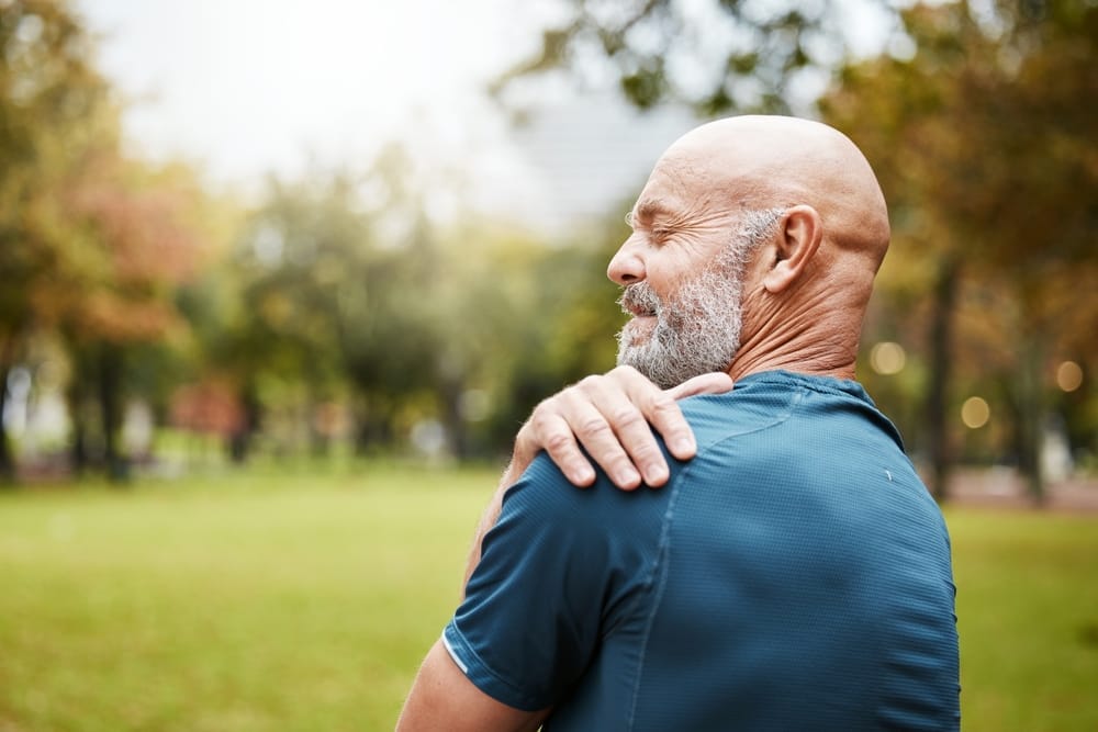 A man outdoors holding his shoulder in discomfort, possibly indicating shoulder pain or injury - Chiropractor Kennesaw GA A man outdoors holding his shoulder in discomfort, possibly indicating shoulder pain or injury - Chiropractor Kennesaw GA